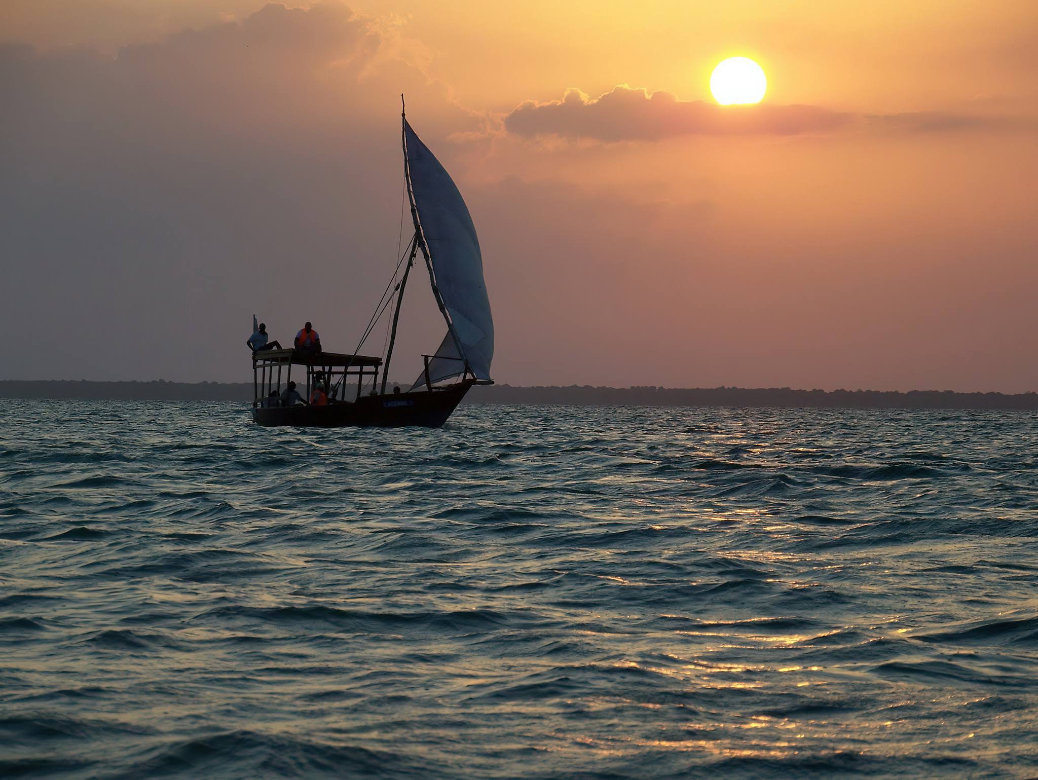 Traditional Zanzibar dhow sailing across calm ocean during colorful sunset with glowing horizon reflections.