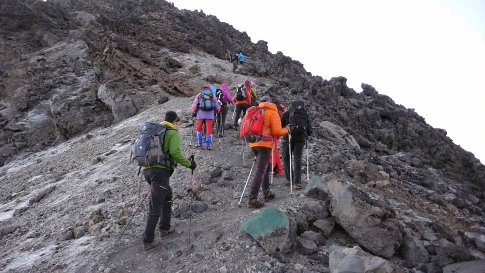 Hikers approaching Mount Meru summit with Kilimanjaro visible above distant morning clouds beyond.