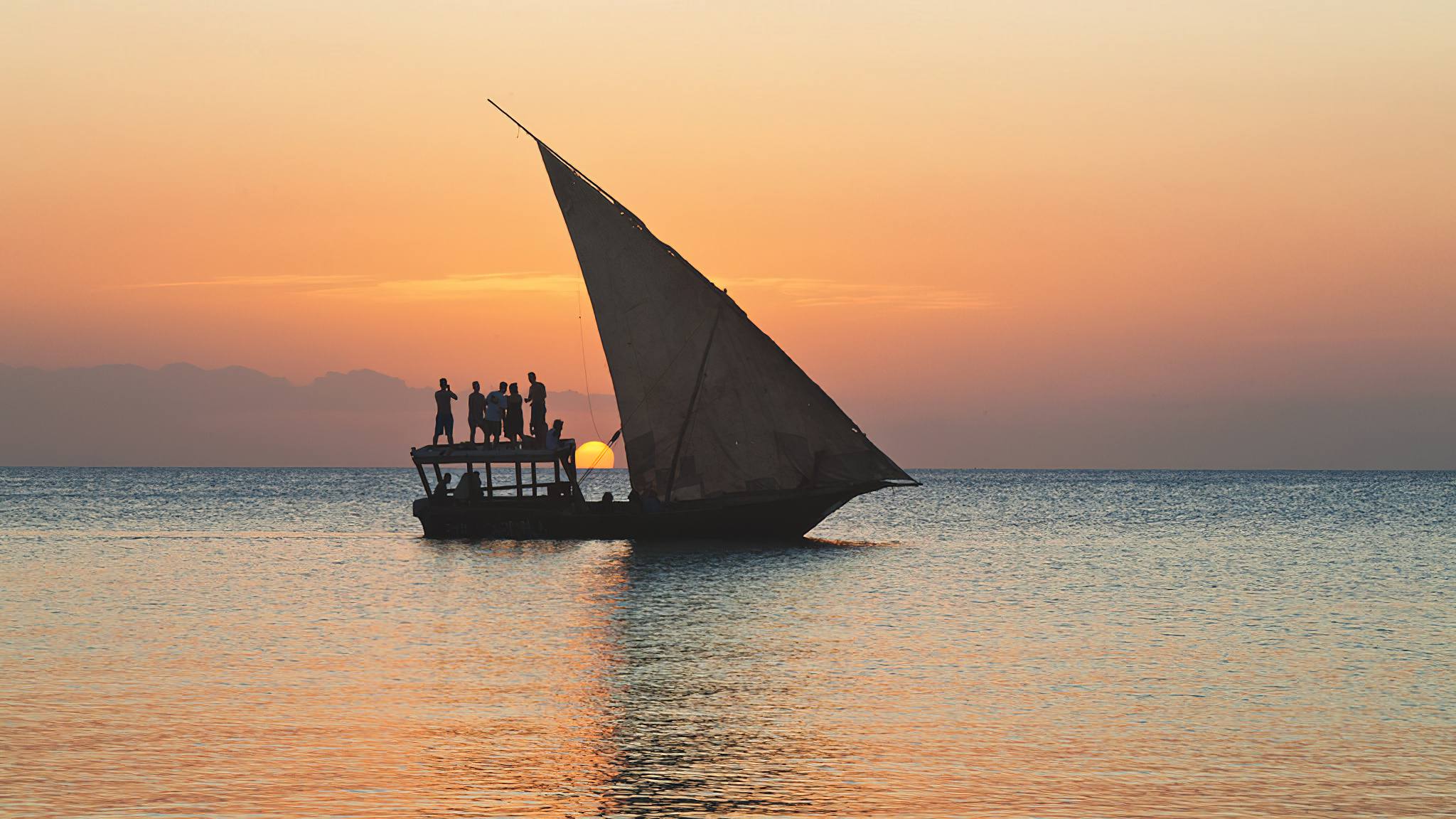 Wooden dhow cruising beside Zanzibar coastline beneath vibrant sunset skies over tranquil Indian Ocean.