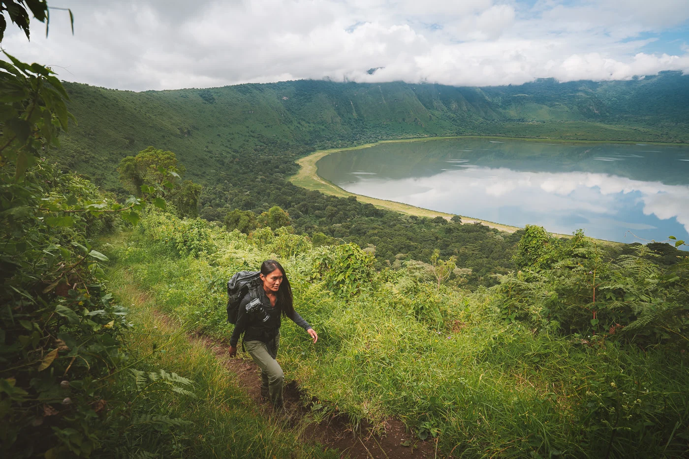 Steep volcanic crater with lake, forest rim, wildlife, and hikers descending trail inside Empakaai.